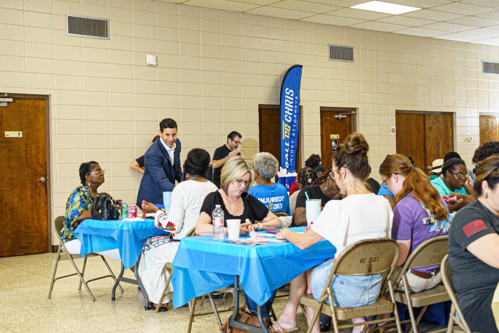 Community event inside a parish hall with several adults seated at round tables covered in blue tablecloths. People are talking, writing, and working on activities together. A man in a suit is leaning in to speak with attendees. A vertical banner in the background reads “Call Chris Injury Attorneys.” The setting appears to be a local gathering or outreach event.